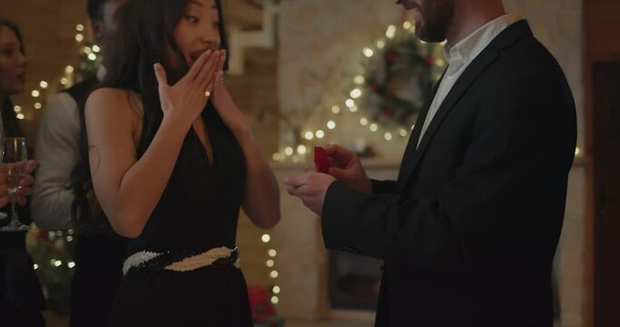 Handsome Boyfriend Proposing, Giving Ring to Beautiful Girl while Having Party with Friends. Romantic courtship. Handsome Young Man Proposing to His Girlfriend while Celebrating New Year Party indoor