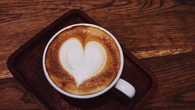 Coffee break and romantic mood concept. Cup of caramel cappuccino with heart shaped foam art made of lactose-free milk, served on wooden table in cafeteria. High quality 4k footage