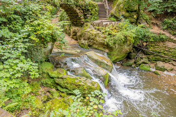 Black Ernz river among wild vegetation, Schiessentümpel waterfall seen from a higher perspective, crystal clear water flowing between rocks, staircase in the background, Mullerthal Trail, Luxembourg
