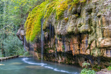 Moss covering a rock formation with crystalline calcareous water falling into a spring, Travertine source Kallektuffquell, trees in the background, Mullerthal Trail, Luxembourg. Long exposure image - obrazy, fototapety, plakaty