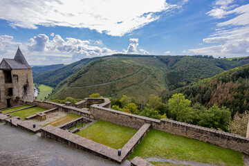 Fototapeta premium Landscape seen from the upper courtyard of the medieval castle of Bourscheid, ramparts, tower and valley, mountains, green trees, dramatic blue sky with abundant clouds in the background, Luxembourg