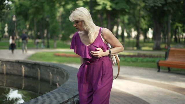 Adult woman walking in park having heart ache sitting down at fountain taking pills. Portrait of sad ill Caucasian lady outdoors. Chronic illness and lifestyle concept