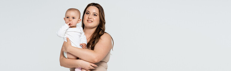 Smiling plus size woman holding baby daughter isolated on grey, banner.