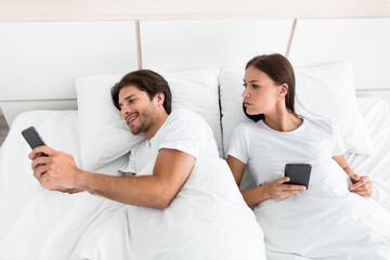 Smiling attractive millennial caucasian man with stubble chats on smartphone, jealous wife looks at phone