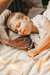 Obraz na płótnie Canvas portrait of a boy sleeping under a white blanket and with a garland. christmas night baby. Little boy sleeping on gray pillow, vertical frame