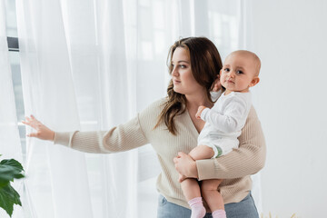Pretty woman with overweight holding baby near curtains at home. - obrazy, fototapety, plakaty