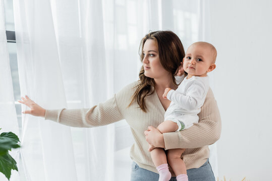Pretty woman with overweight holding baby near curtains at home.