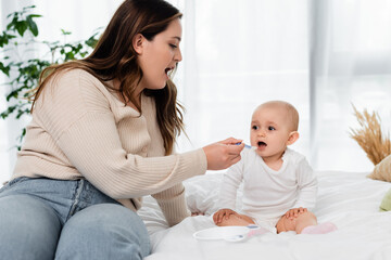 Plus size mother open mouth while feeding baby on bed.