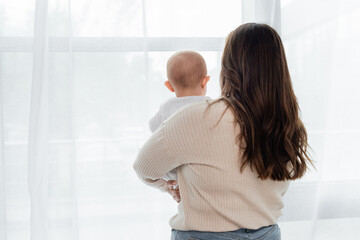 Back view of plus size woman holding baby near window at home.