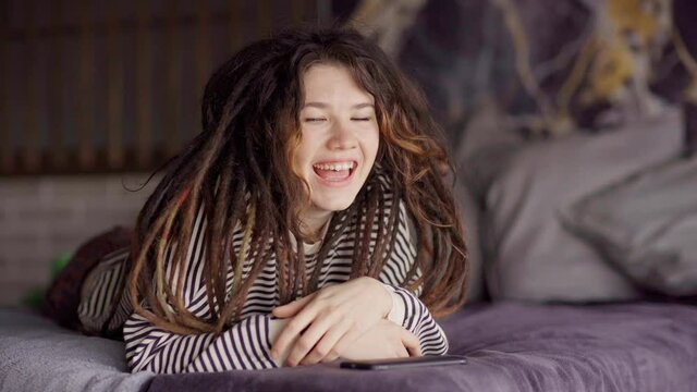 Portrait of smiling playful young woman with dreadlocks and pierced tongue looking at camera lying on bed at home with smartphone. Cheerful girl enjoying relaxing in bedroom