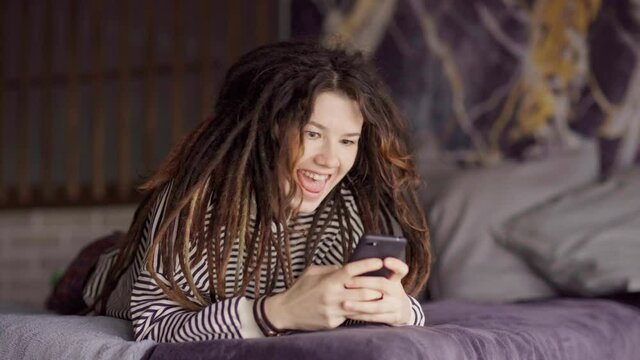 Smiling young woman with dreadlocks and tongue piercing having fun text messaging on cell phone lying on bed at home. Cheerful girl laughing while typing sms on smartphone in domestic bedroom