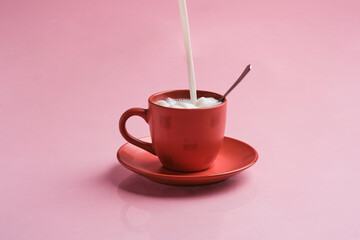 red mug on a pink background with reflection. Pouring milk into the cup with a spoon inside