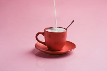red mug on a pink background with reflection. Pouring milk into the cup with a spoon inside