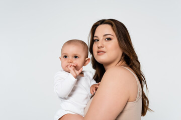 Pretty body positive woman hugging baby girl and looking at camera isolated on grey.