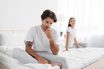 Depressed millennial european husband with stubble sits on bed and sad, wife apologizes in bedroom - obrazy, fototapety, plakaty
