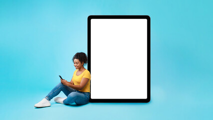 Young black lady using cellphone, sitting next to giant tablet pc with empty white screen, mockup for app or website