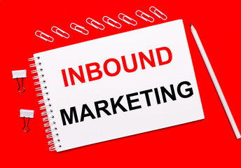 On a bright red background, a white pencil, white paper clips, and a white notebook with the text INBOUND MARKETING