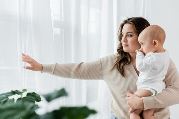 Young body positive mother holding child near curtains at home.