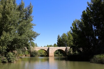 Fototapeta na wymiar Roman bridge over the River Pisuerga seen from the river bank on a hot summer day Lantadilla Palencia Spain