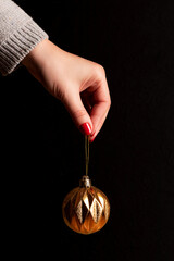 Female hand in gray knitted sweater with golden Christmas ball with beautiful manicure - red nails against black background. Nail and skin care concept