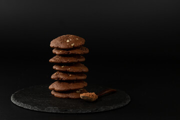 chocolate cookies are stacked on a black stone board on a black background, next to a wooden spoon with cocoa