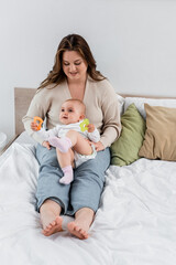 Positive plus size mother looking at baby with toys on bed.