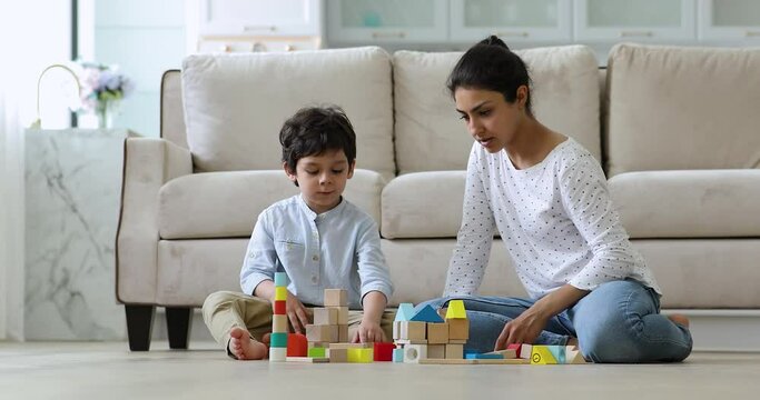 Playtime. Focused mom nanny babysitter sit on heated floor with preschool age boy play creative game. Indian adult elder sister kindergarten teacher help kid in building castle of bright colored brick