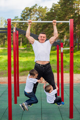 Fototapeta premium Caucasian man and two boys doing exercises outdoors. The father pulls himself up on the horizontal bar with his sons on the playground.