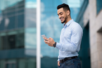Positive middle eastern guy standing by business center, using smartphone