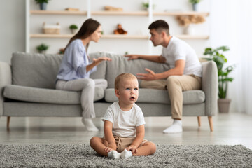 Family Conflicts. Little Baby Boy Sitting Alone While Parents Quarreling On Background