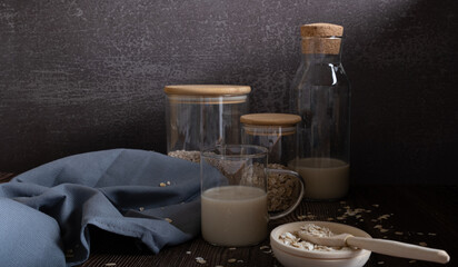 Oat milk in a glass glass with cans of oat flakes and grain on a dark background. Low key