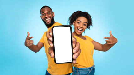 Joyful African American couple pointing at smartphone with empty white screen, advertising your app...
