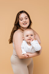 Smiling plus size mom holding positive baby daughter isolated on beige.