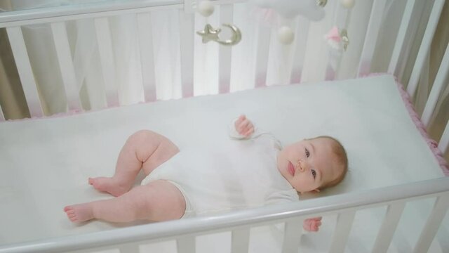 Beautiful baby girl relaxing on her baby cot alone looking up at the toys cot and playing with hands very cute. 4k