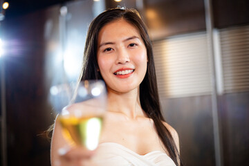 Portrait of attractive beautiful Asian woman holding glasses of wine or beer alcohol toasting to camera, focus on female happy smile face, lifestyle party music with light at night on rooftop 