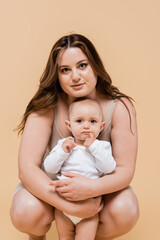 Young plus size woman looking at camera while hugging child isolated on beige.