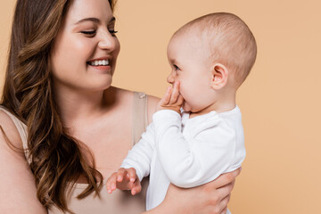 Cheerful plus size mother looking at baby daughter isolated on beige.