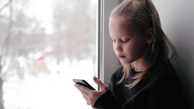 Little blonde girl in black sweater sitting near window and using smartphone after school. Bored child writing on phone. Sad kid home alone during lockdown