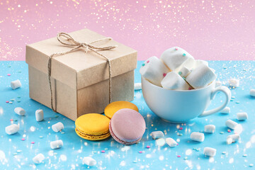 Colorful macaroons, gift box and a cup of hot chocolate decorated with marshmallows on a blue background. Sweet desserts. Gift for your holiday.
