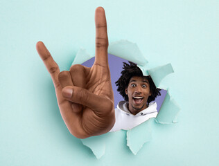 Excited black teenager showing huge rock gesture through hole in light blue ripped paper