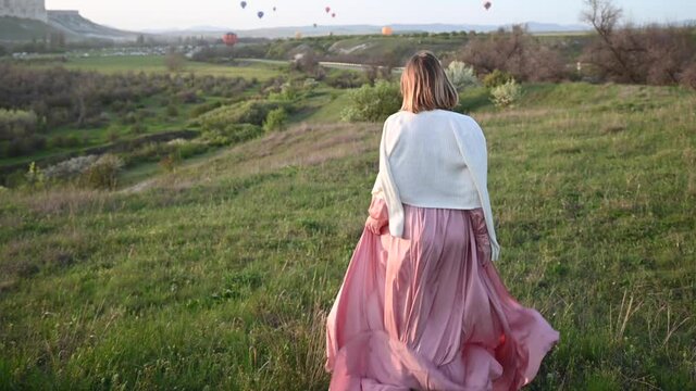 Red haired thin beautiful young girl in a pink long dress fluttering in the wind and a white jacket falling from her shoulders runs down the slope to the colorful balloons
