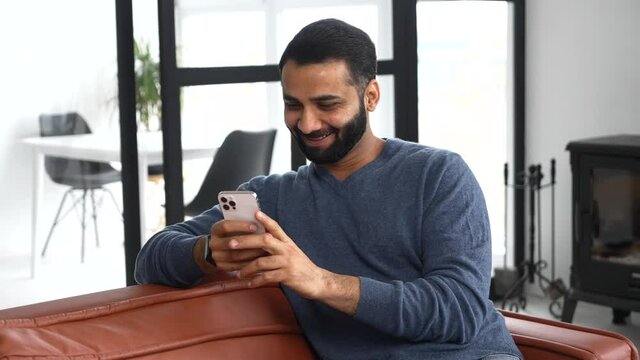 Cheerful Indian guy wearing casual outfit messaging, texting on the smartphone. Multiracial bearded man sits on the modern sofa and using mobile app, scrolling feed news, websurfing, playing game