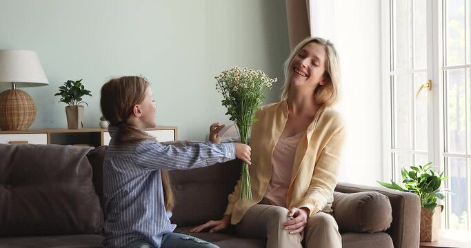 Young female sit on couch with closed eyes before receive March 8 surprise from caring daughter. Happy little girl greet grown elder sister mommy on Mothers Day hug give flowers card with best wishes
