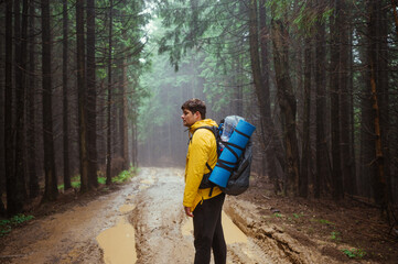 Hiker man with a backpack and a yellow jacket stands on the road in a mountain forest and looks to the side against the backdrop of a beautiful misty landscape.