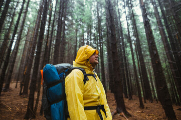 Young male hiker in a raincoat stands in the woods and looks away with a serious face on a background of beautiful view