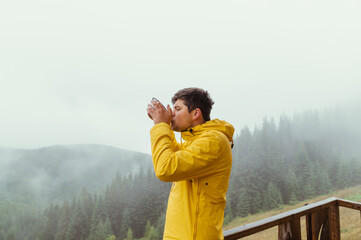 Male hiker in a yellow jacket drinks soup from a plate in the mountains on a background of beautiful views of the cloudy forest.