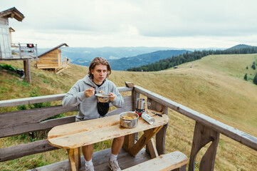 Naklejka na ściany i meble Young man eating pasta for breakfast on the terrace of a wooden house in the mountains. Tourist eating in the mountains