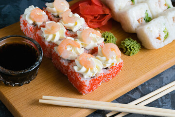 Appetizing and delicious sushi rolls with shrimp, cheese, caviar, chicken, soy sauce, nori, wasabi and ginger on a wooden board on a dark background