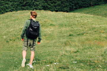 Fototapeta premium Young man with a backpack on a hike walks on a meadow in the mountains and looks away, copy space on the right. A tourist walks in the mountains.