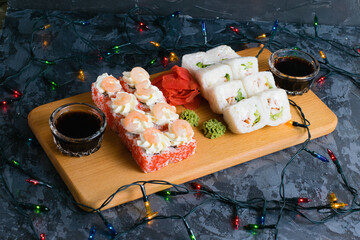 Appetizing and delicious sushi rolls with shrimp, cheese, caviar, chicken, soy sauce, nori, wasabi and ginger on a wooden board on a dark background with a Christmas garland